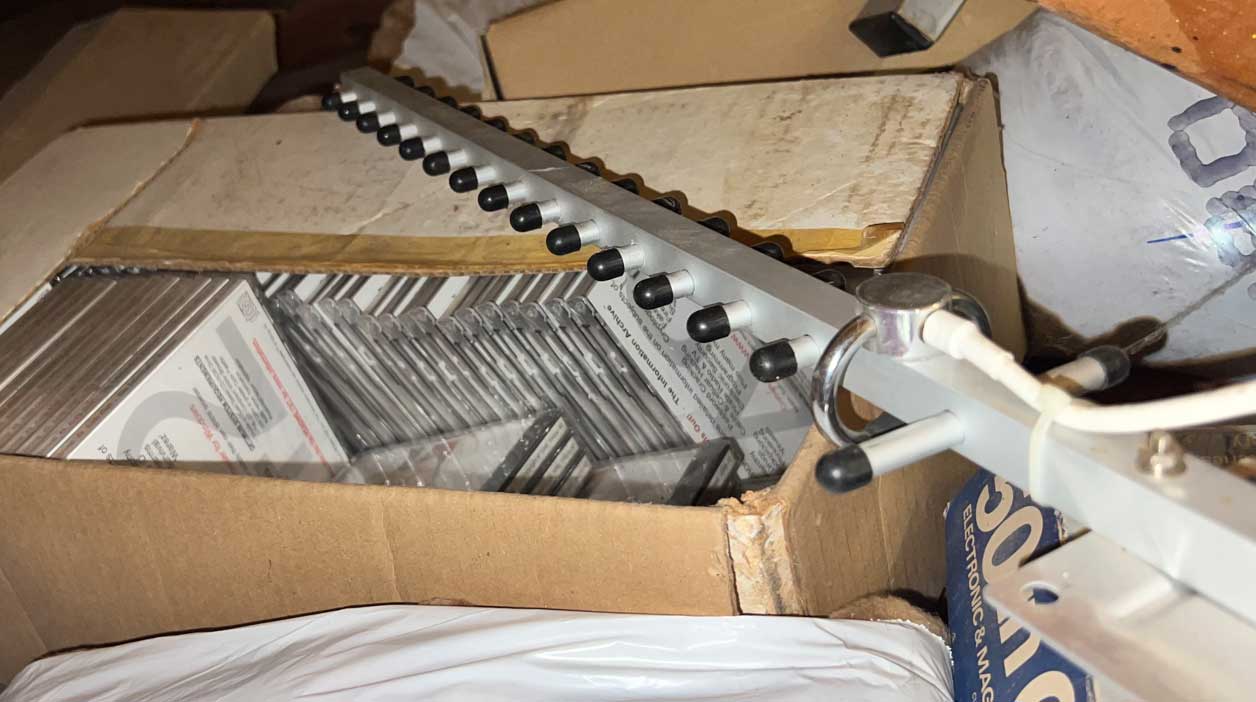 Open cardboard box filled with stacks of jewel-case CDs and printed inserts, stored in an attic or storage space alongside packaging materials and a 900 MHz Yagi antenna.