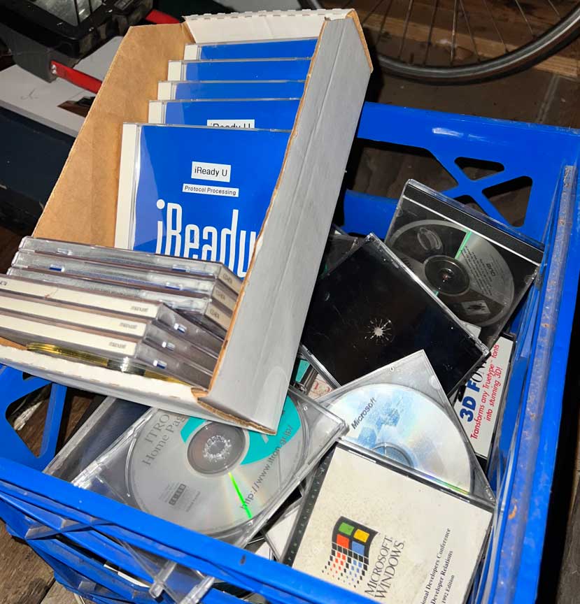 Box of vintage software CDs and manuals, including iReady U protocol processing binders and Microsoft Windows discs, stacked inside a blue plastic crate in a workshop setting