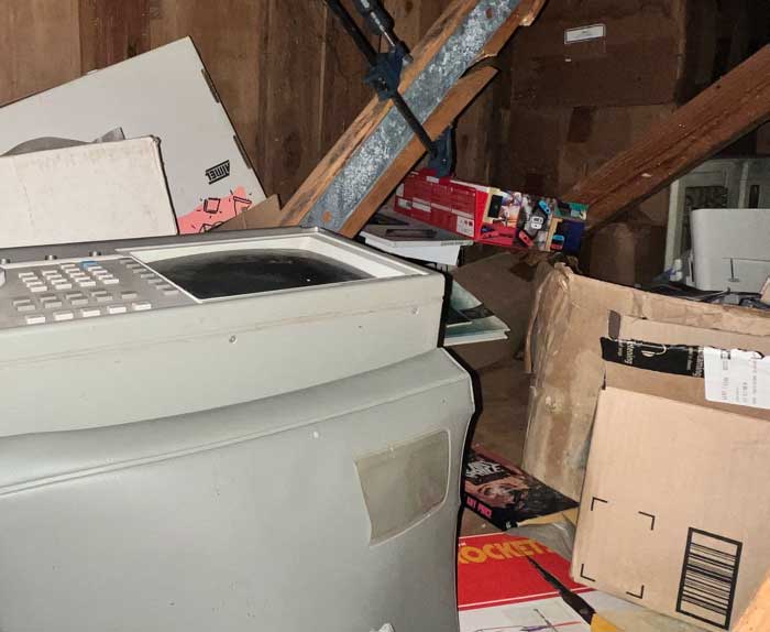 Dusty attic storage space with an old beige office machine, cardboard boxes, and scattered 1990s computer and electronics packaging among exposed rafters.