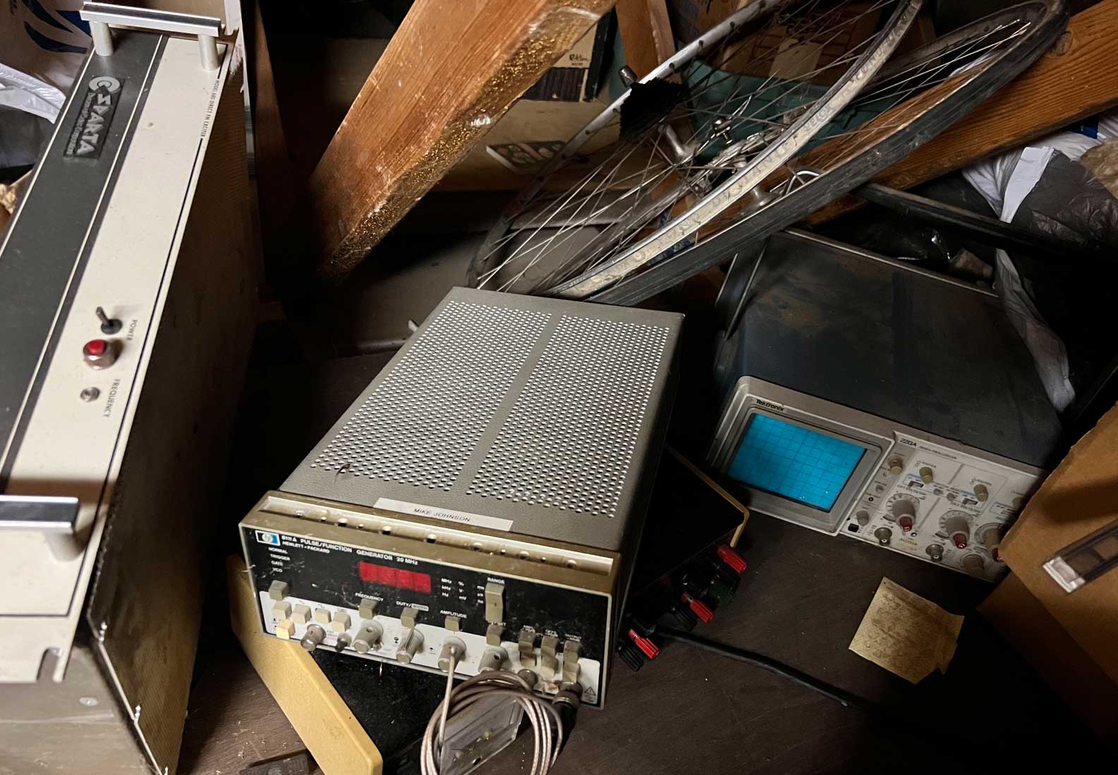 Dusty attic storage area with vintage electronics test equipment, including an HP signal generator and a Tektronix oscilloscope, surrounded by boxes, wood beams, and a bicycle wheel.