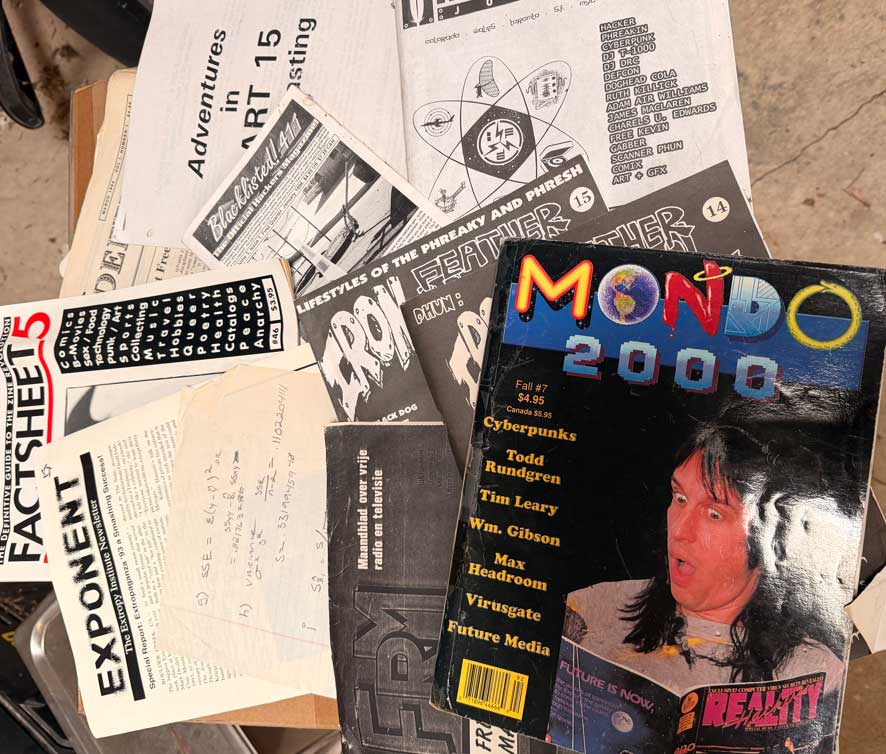 "Open cardboard box on a concrete floor containing 1990s cyberculture magazines and zines, including Mondo 2000, 2600, Iron Feather Journal, and handwritten notes on lined paper.”