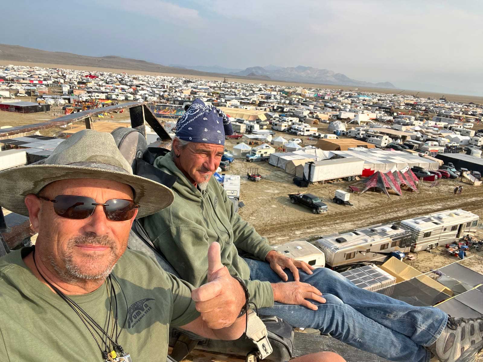 Camp members riding a couch strapped to a telehandler, lifted high above Burning Man to survey storm damage across Black Rock City after heavy rain and wind.