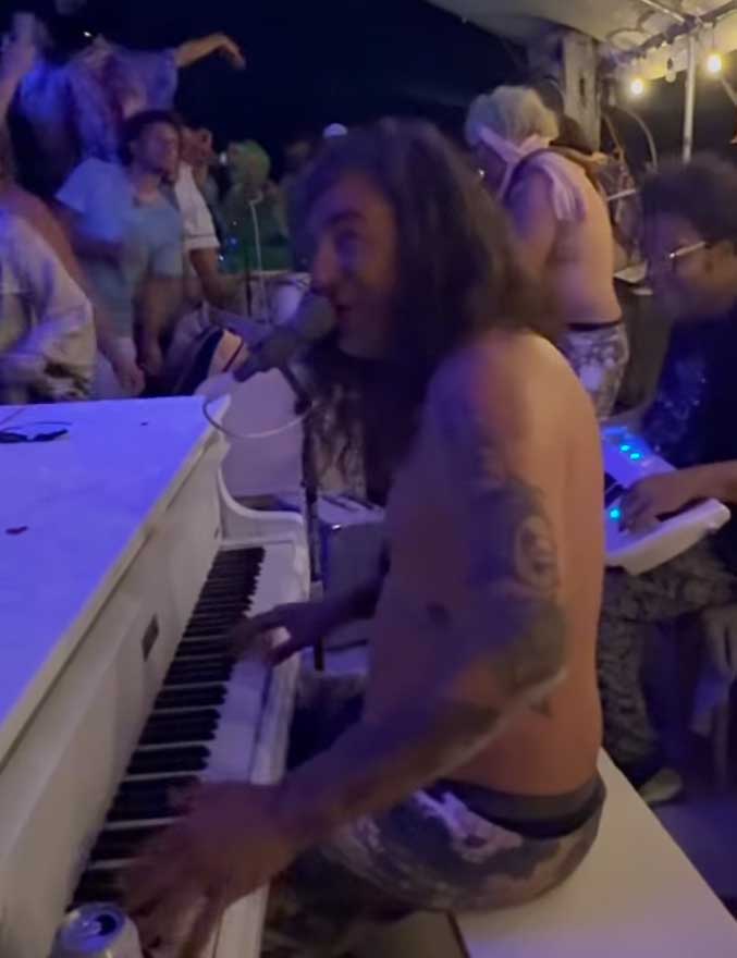 Milton John performing shirtless at a white piano in the Black Rock Piano Lounge at Burning Man, with Burners dancing around.