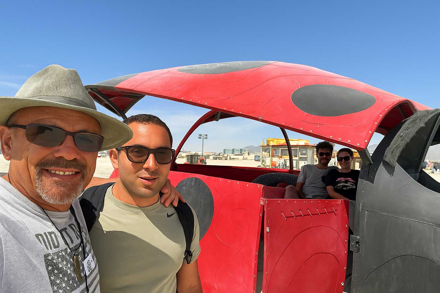 Father and son stand beside the red Ladybug art car at Burning Man, smiling before heading to Burner Express before the Temple burn.