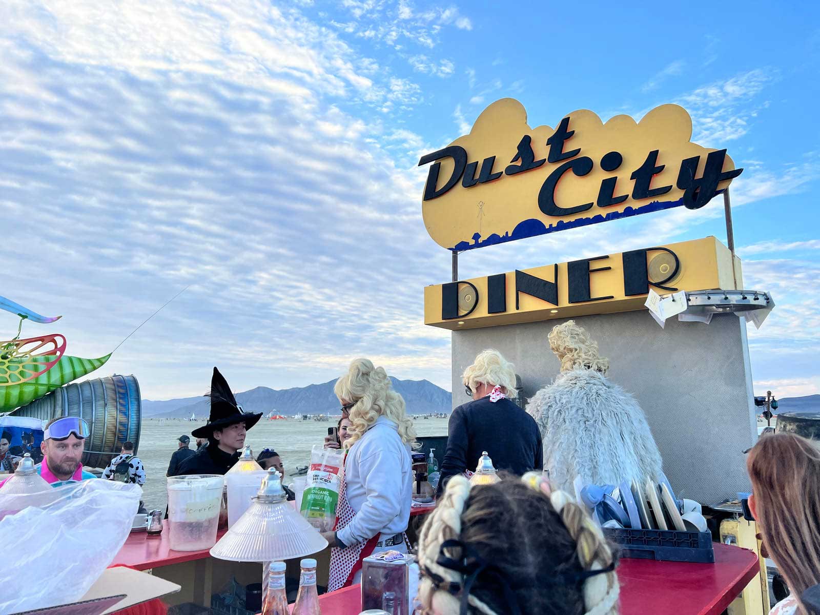 Dust City Diner pop-up in deep playa at Burning Man, styled as a 1950s diner with stools, counters, and neon glow.