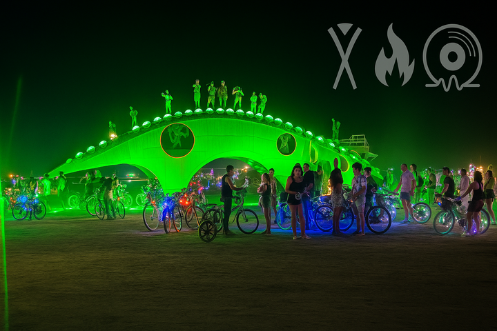 Guma, the glowing steel dinosaur art piece at Burning Man, lit in green at night and surrounded by bikes and people.