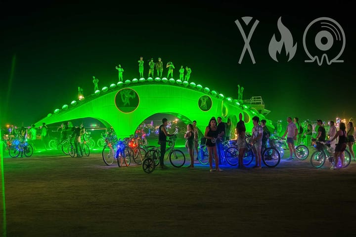 Guma, the glowing steel dinosaur art piece at Burning Man, lit in green at night and surrounded by bikes and people.